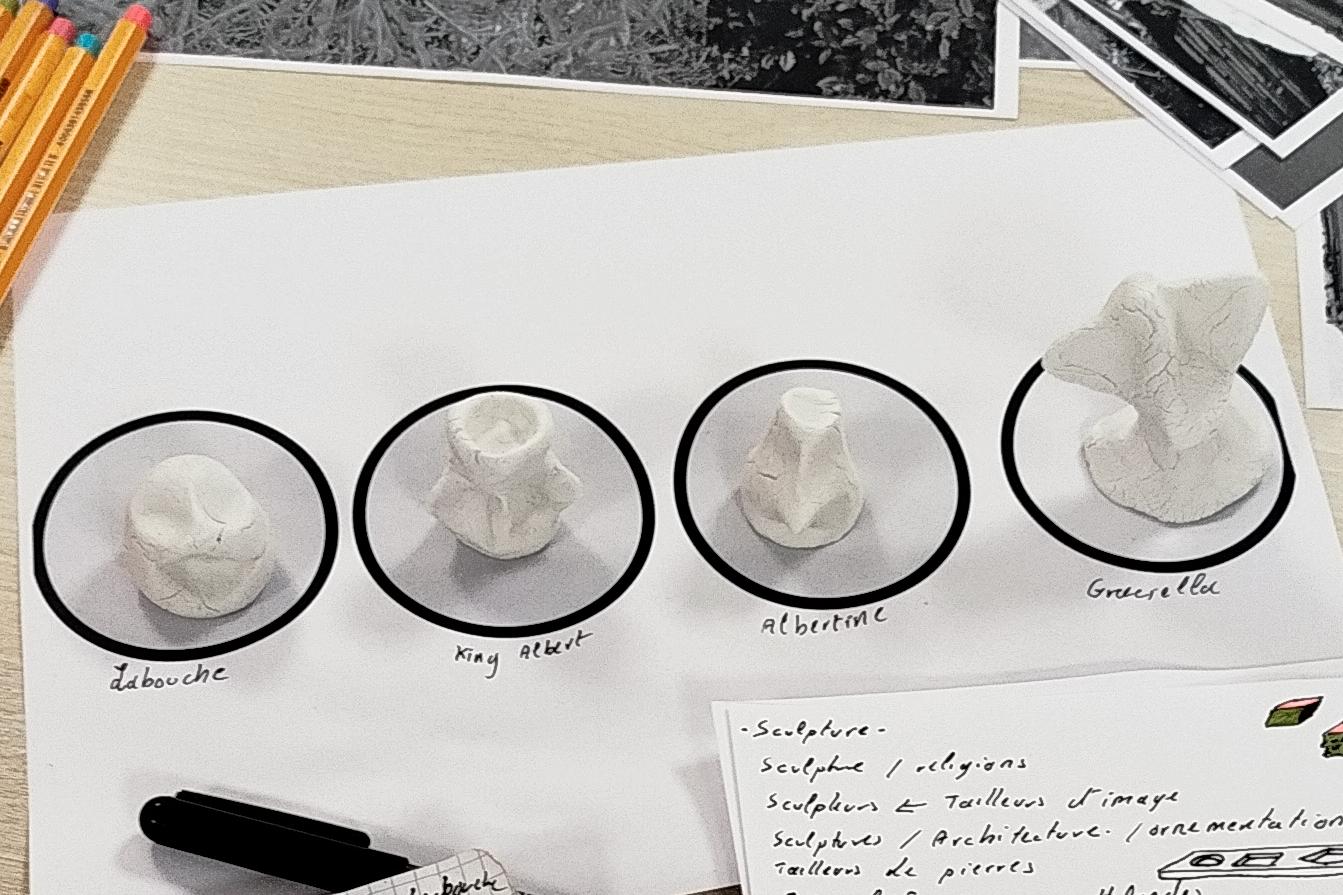 Posés sur une table, des feutres, des photos de sculptures, 4 personnages en pâte à modeler et des notes prises sur des feuilles et sur un carnet